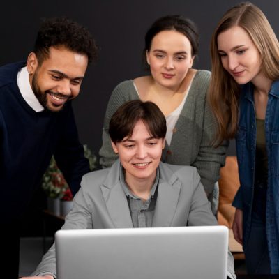 Four people around a laptop, one seated with a coffee mug, others standing.